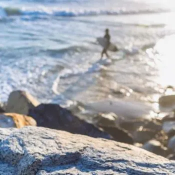 A bottle of white wine sits on a rock near the ocean, with two people walking in the water and waves in the background.
