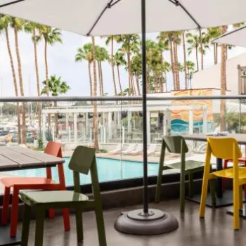 Outdoor dining area with colorful chairs and tables under umbrellas, overlooking a pool, palm trees, and marina with boats in the background.
