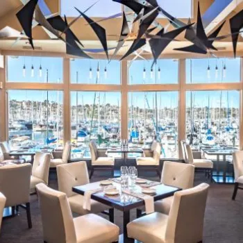 Modern restaurant dining area with white chairs and set tables, large windows overlooking a marina with numerous docked boats in the background.