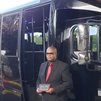 A man in a black suit and red tie holds a plaque while standing in front of a large black bus parked on a street lined with trees and plants.