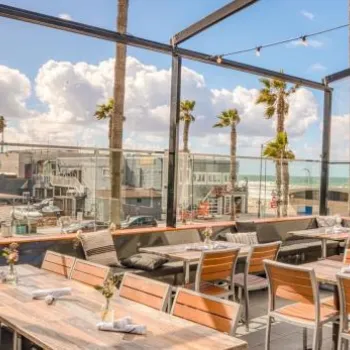 Outdoor restaurant patio with wooden tables and chairs overlooks a beachside street, palm trees, and ocean under a partly cloudy sky.