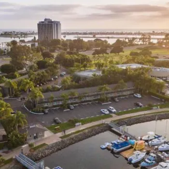 Aerial view of a marina with boats docked, adjacent buildings, palm trees, a high-rise hotel, and water in the background at sunset.