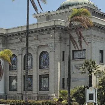 A large, historic building with columns, arched windows, stained glass, palm trees, and a dome on the roof under a clear sky.