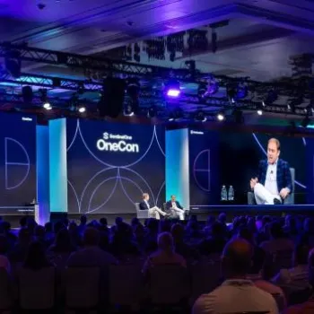 A speaker sits on stage in front of a large audience at a conference, with two large screens displaying the event branding and the speaker’s image.
