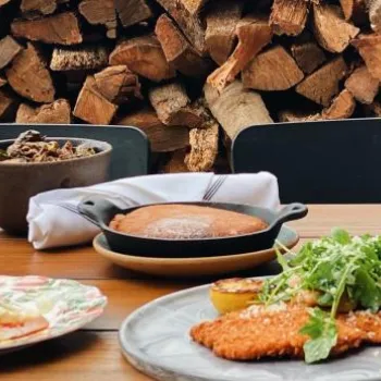 A wooden table set with various plated dishes, including meats and salads, in front of a stacked firewood backdrop.