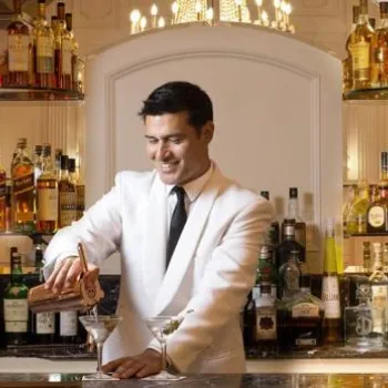 A bartender in a white jacket pours a drink into a martini glass behind a well-stocked bar with various bottles and glassware.