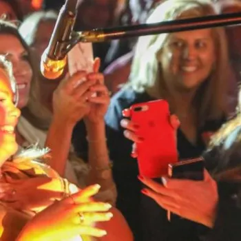 A group of people smiles and takes photos with their phones as a woman stands under a microphone, brightly lit, at an event.