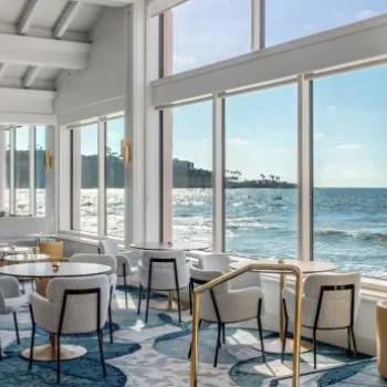 Bright, modern dining area with large windows overlooking the ocean; sunlight streams in, illuminating light-colored furniture and wood floors.