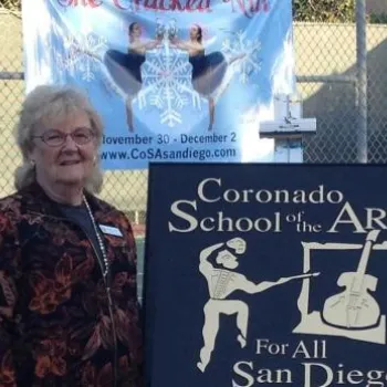 Four adults stand outdoors next to a "Coronado School of the Arts For All San Diego" sign with a fence and event banners in the background.