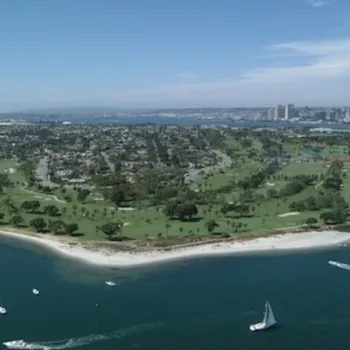 Aerial view of a peninsula with green spaces, residential areas, surrounding water with boats, and a city skyline in the background under a blue sky.