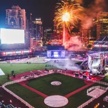 A nighttime stadium scene with fireworks, bright stage lights, and a partially filled audience, set against a city skyline.