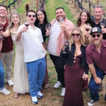 A group of thirteen adults standing outdoors in a vineyard, holding wine glasses and smiling at the camera.