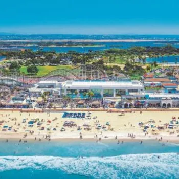 Aerial view of a crowded beach with waves, a boardwalk, an amusement park with a roller coaster, parking lots, and surrounding greenery under a clear blue sky.