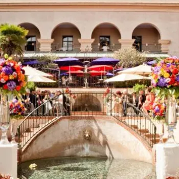 Elegant outdoor event with colorful flower arrangements, guests seated around a fountain, and a large building with arches in the background.
