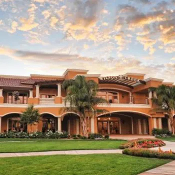 A large, elegant building with arched entrances and balconies is surrounded by green lawns, palm trees, and landscaped gardens under a partly cloudy sky at sunset.
