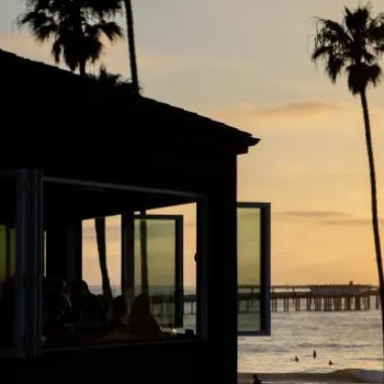 A beachside building with open windows is silhouetted against a sunset, with palm trees and a pier visible in the background over the ocean.