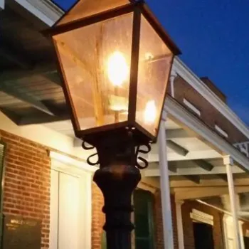 A lit gas lantern is mounted in front of a brick building with green shutters and a covered porch at dusk.