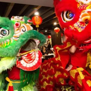 Colorful lion dancers perform indoors at a festive event, with red lanterns hanging from the ceiling and people watching in the background.