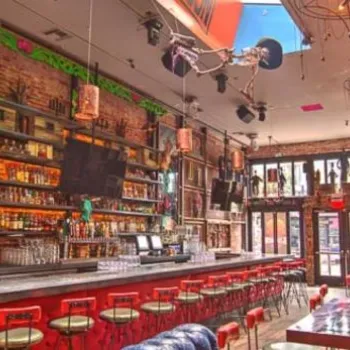 Colorful bar interior with a long counter, bar stools, high ceilings, exposed brick walls, large windows, and eclectic wall art and lighting fixtures.