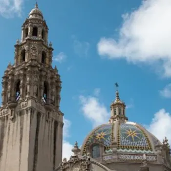 Ornate tower and dome of a historic building with mosaic details, palm trees, and a partly cloudy sky in the background.
