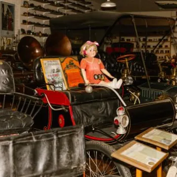 A vintage automobile display in a museum room, featuring an old car with a doll seated inside and various antiques and collectibles visible in the background.