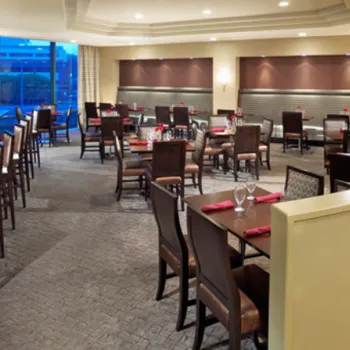 Modern restaurant interior with tables and chairs neatly arranged, a bar area on the left, and large windows letting in natural light. A woman stands behind the bar.