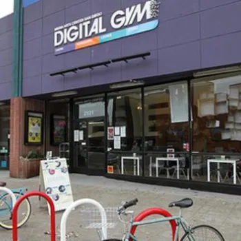 Storefront of Digital Gym, featuring large windows, a purple sign, bike racks with bicycles, and various posters displayed in the windows.