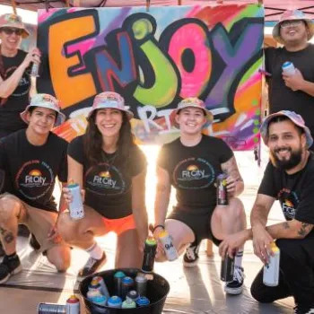 A group of people in matching outfits pose in front of a colorful "Enjoy" graffiti banner, holding spray paint cans, at an outdoor event.