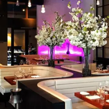 Modern restaurant interior with white booth seating, wooden tables, elegant place settings, and large vases of white flowers; purple accent lighting along a brick wall.