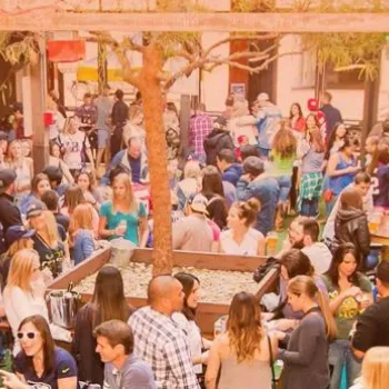 Large crowd of people gathered in an outdoor courtyard, socializing and sitting at tables under partial shade.