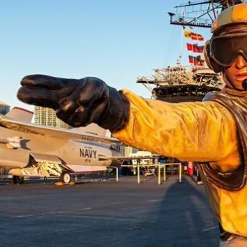 A flight deck crew member in a yellow jacket signals on an aircraft carrier with fighter jets and ship structures visible in the background.