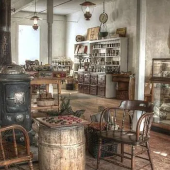 Vintage general store interior with wooden floors, shelves of goods, a potbelly stove, wicker chairs, and a glass display case. Light streams in from a window on the right.