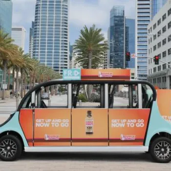 A small electric shuttle with "GET UP AND GO NOW TO GO" advertisements on its side is parked in front of tall buildings and palm trees in an urban area.