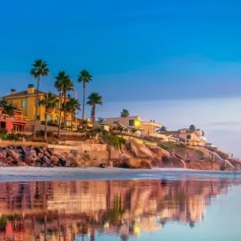 Beachfront houses and palm trees reflected on wet sand during sunset, with a calm ocean and a colorful sky in the background.