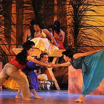 Five dancers perform on stage, two balancing in front while three sit or recline in the background; the set includes tropical plants and warm lighting.