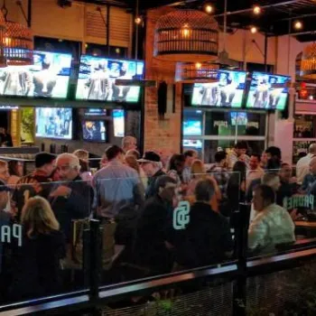 A crowded sports bar at night with people seated at tables and the bar, multiple TVs displaying games, and neon signs visible in the background.