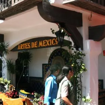 Two people walk past colorful outdoor decorations and a store with a sign reading "Artes de Mexico" on a lively street with umbrellas and vibrant flowers.