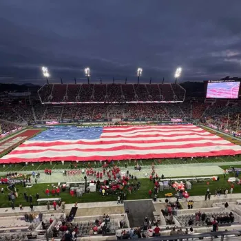 A large American flag is unfurled across a football field before a game, with players, performers, and a crowd in the stadium under stadium lights.