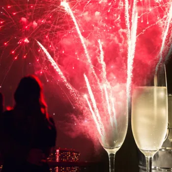 Two people watch red fireworks in the night sky, with two champagne glasses and an ice bucket in the foreground.