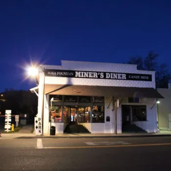 A white building at night with a sign reading "Miner's Diner" above the entrance. Lights illuminate the soda fountain and candy shop inside. A "PARK" sign stands at the corner.