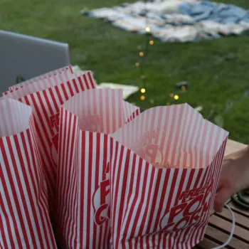 Four striped popcorn bags sit on a wooden table outdoors, with a person's hand nearby and picnic blankets and string lights visible on the grass in the background.