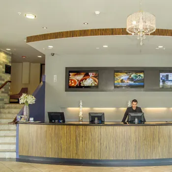 A hotel reception area with a curved wooden front desk, a staff member behind the counter, and three display screens on the wall above.