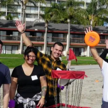 A group of people on a beach stand around a disc golf basket, holding frisbees and raising their arms, with palm trees and buildings in the background.