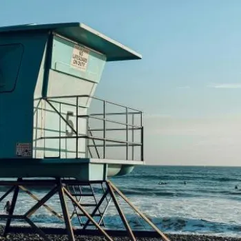 A blue lifeguard tower stands on a pebbled beach overlooking the ocean with a few surfers in the water under a clear sky.