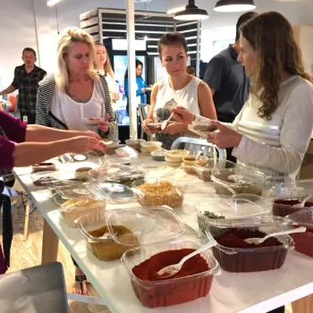 Several people stand around a table with open containers of spices and dried foods, sampling and discussing the contents in a brightly lit room.