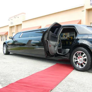 A black stretch limousine with an open rear door is parked on a street; a red carpet is rolled out beside it toward a building.