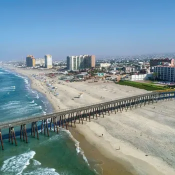 A long wooden pier extends over the ocean from a sandy beach, with waves breaking along the shore and tall buildings lining the coastline under a clear blue sky.