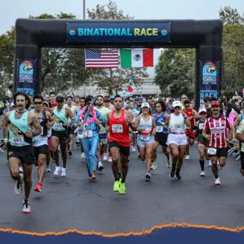 A group of runners start a race under an archway labeled "Binational Race," with U.S. and Mexican flags above and spectators on both sides of the course.