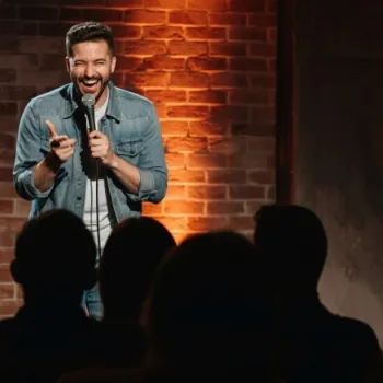 A man in a denim jacket stands on stage performing stand-up comedy, smiling and holding a microphone, with an audience seated in front of him.