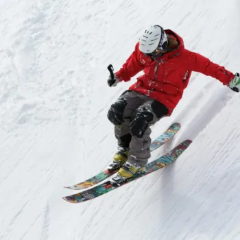 A skier in a red jacket and white helmet is captured mid-air while skiing downhill on a snowy slope.